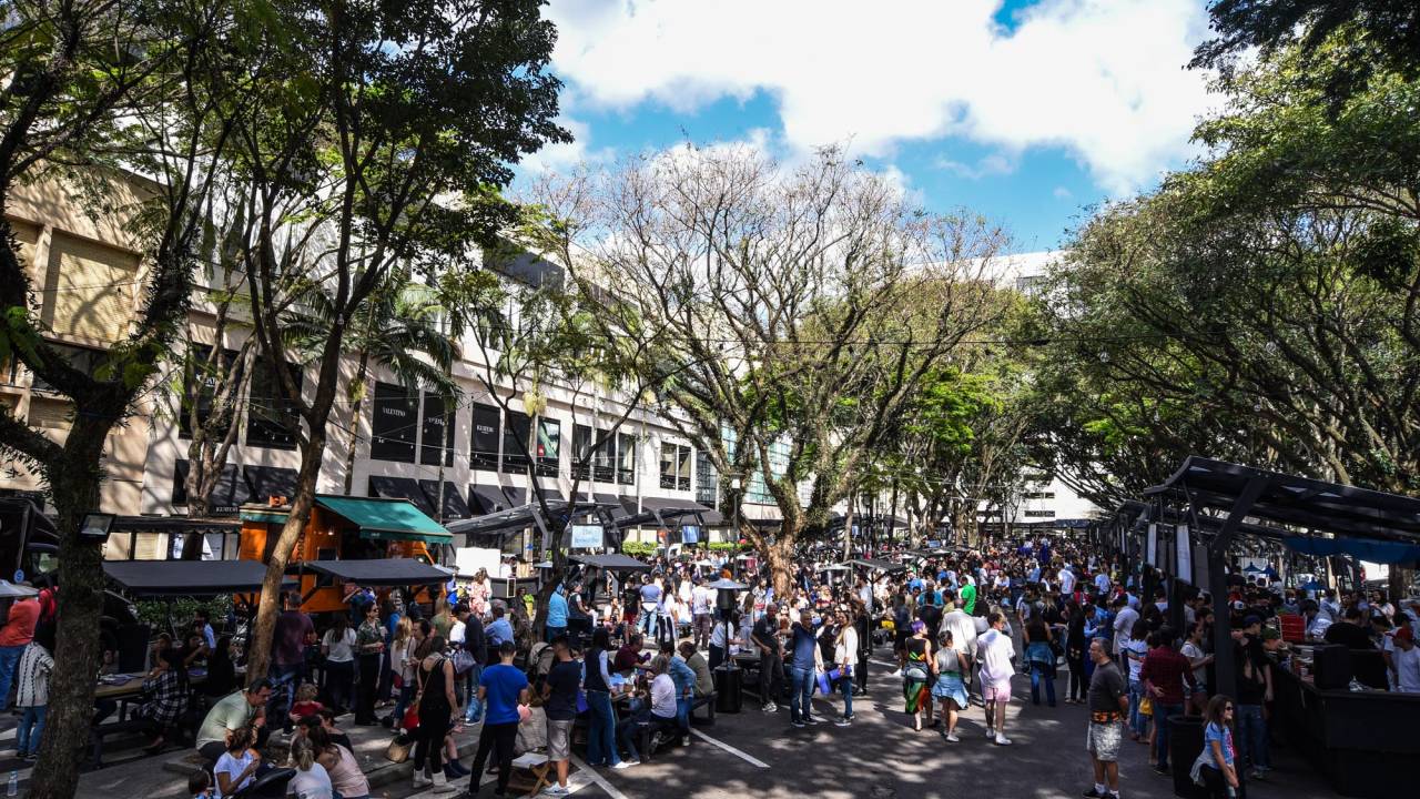 Foto de feira ao ar livre lotada de pessoas com árvores ao redor.