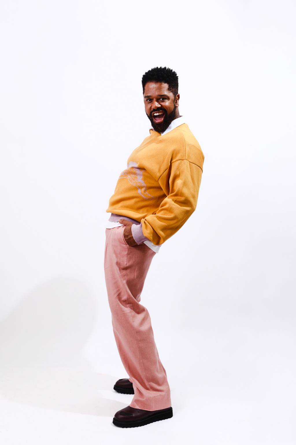Homem negro posa de pé, de lado, com o quadril levemente curvado. Está com as mãos no bolso e veste suéter laranja com calça rosa.