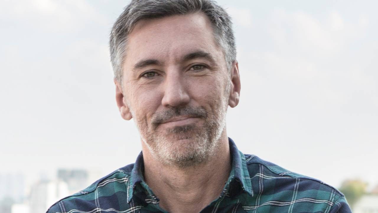 Homem branco grisalho posa de braços cruzados vestindo camisa longa quadriculada nas cores preto e verde-escuro.