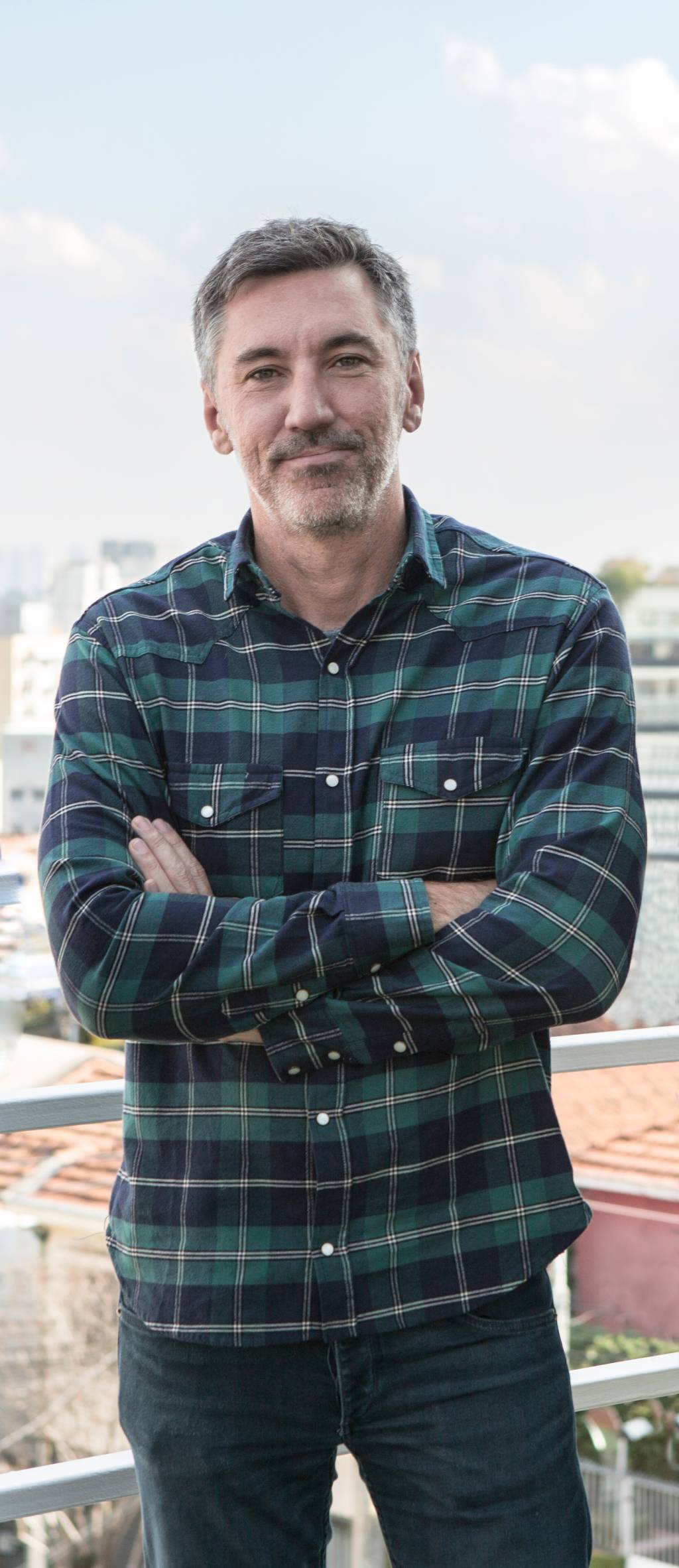 Felipe Cama (Divulgação) Homem branco grisalho posa de braços cruzados vestindo camisa longa quadriculada nas cores preto e verde-escuro.