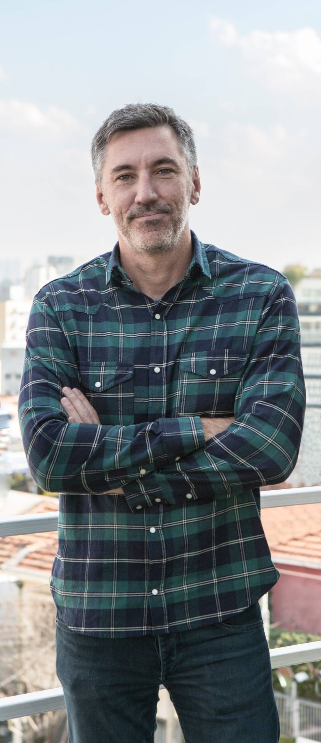 Homem branco grisalho posa de braços cruzados vestindo camisa longa quadriculada nas cores preto e verde-escuro.