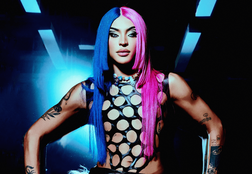 Drag queen posa com peruca dividida em duas cores, azul e rosa, e veste peça preta com furos grandes. Ela exibe seus braços tatuados com as mãos próximas à cintura.