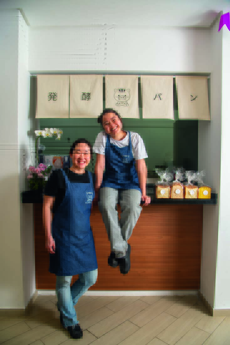 Duas mulheres asiáticas, uma sentada sobre balcão de padaria e a outra em pé ao lado.