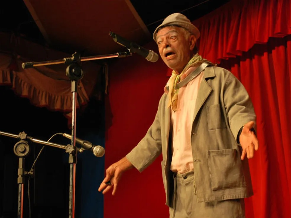 Foto de palhaço falando em microfone com cortina vermelha nos fundos.