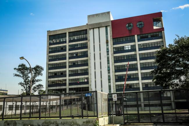 Imagem mostra fachada de edifício, com portões gradeados, gramado e prédio ao fundo, com oito andares