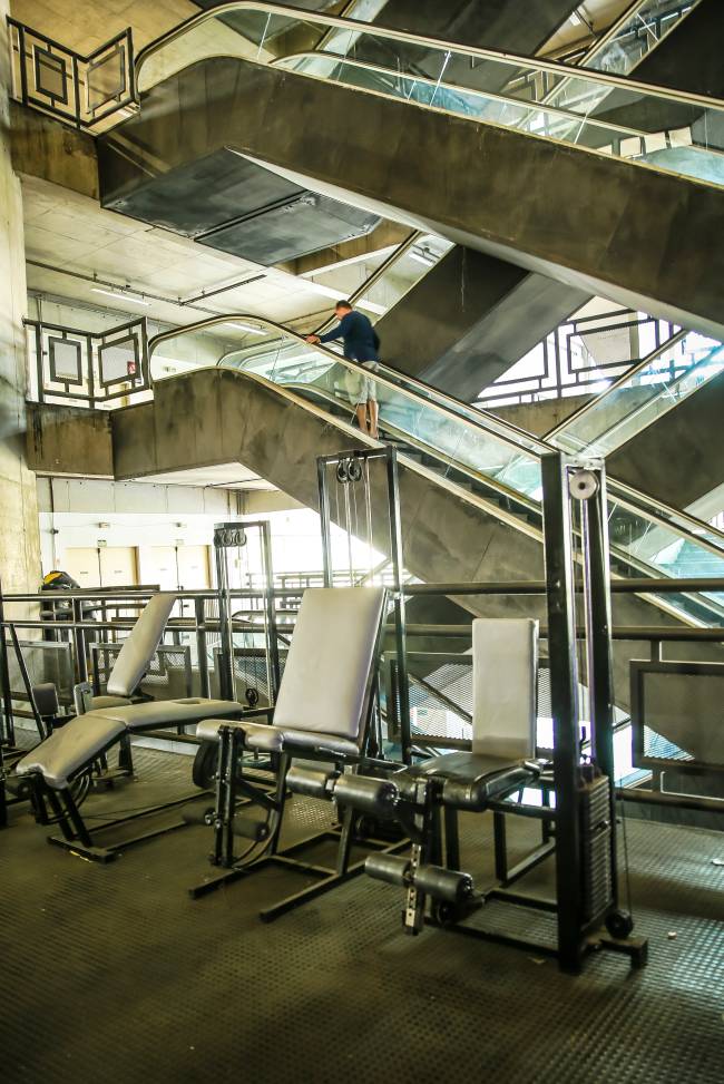Imagem mostra 3 equipamentos de academia e, na parte de trás, escadas rolantes no interior de edifício
