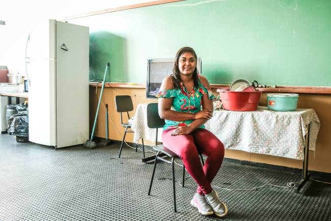 Imagem mostra mulher sentada em cadeira em sala de aula. Na parte de trás, em frente a uma lousa, uma geladeira, vassoura e uma mesa com utensílio de cozinha