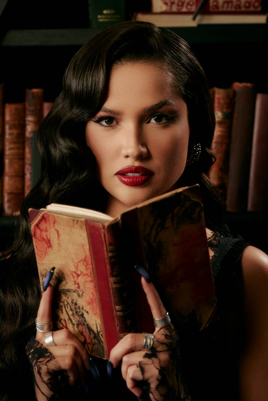 Mulher branca segura livro antigo com dedos indicadores em destaque. Tem cabelos pretos levemente encaracolados e batom bem vermelho.