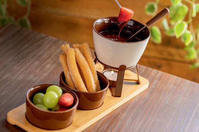 Mesa de madeira com tábua de madeira com mini panela de fondue de chocolate ao lado de potinhos com frutas e churros.