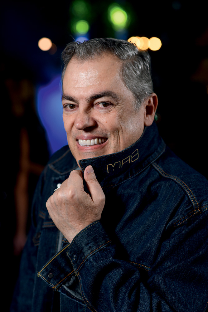 Homem grisalho posa sorrindo e cobrindo o pescoço com a gola da jaqueta jeans escura.