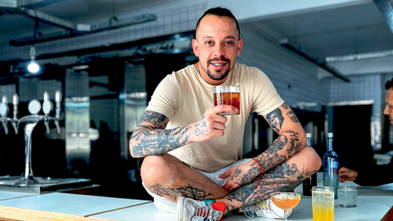 Gabriel Santana, dono do bar, está sentado em um balcão segurando um copo com drinque.