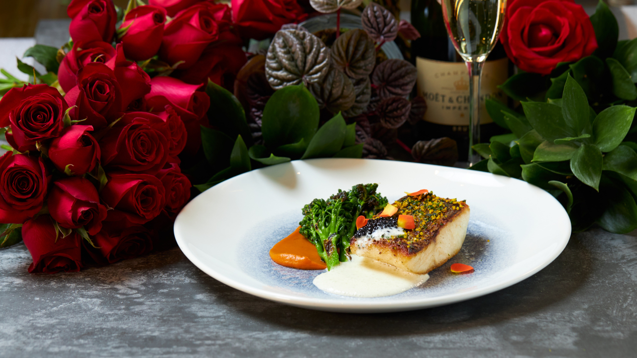Prato de louça branca em frente a bouquet de rosas vermelhas e taça de champanhe com pedaço de pescado, molho branco e brocólis.