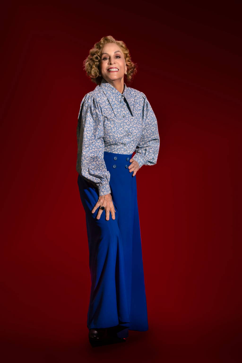 Senhora branca de cabelos grisalhos posa em fundo vermelho vestindo saia azul escuro e camisa azul claro. Ela sorri e apoia uma das mãos na perna.