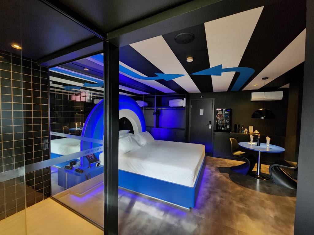 Imagem de quarto de motel em tom azul e repleto de luzes neon, com cama de casal e chuveiro à esquerda. No teto, faixas em preto e branco com grandes setas azuis