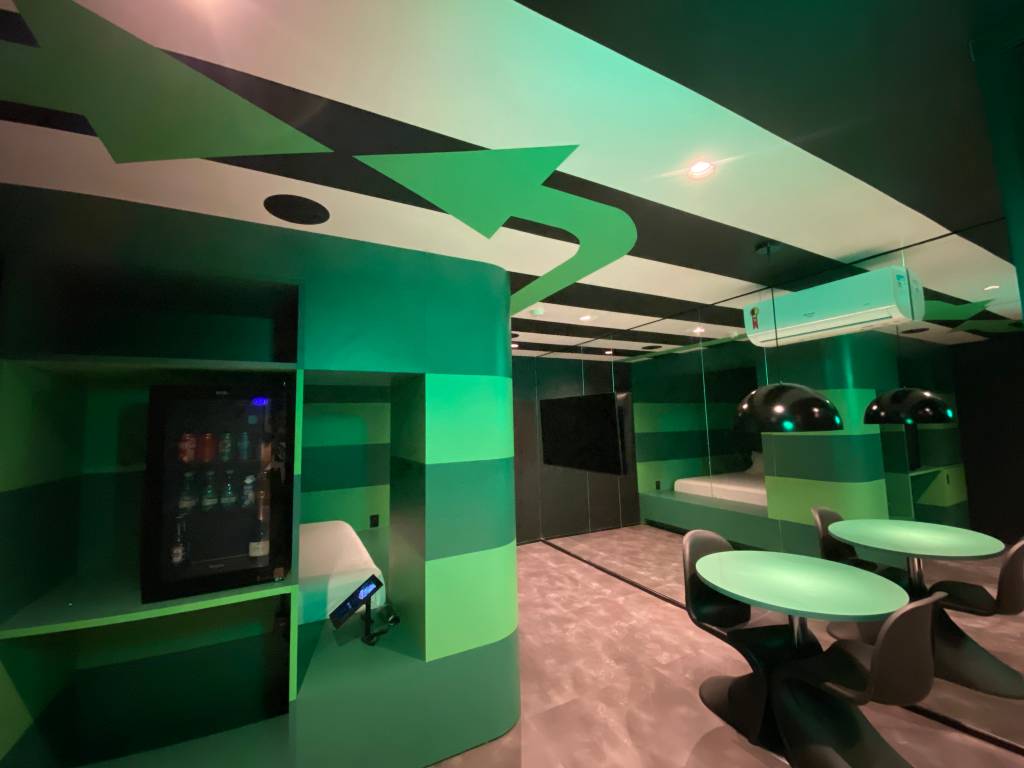 Imagem de quarto de motel em tom verde e repleto de luzes neon, com cama de casal e chuveiro à esquerda. No teto, faixas em preto e branco com grandes setas verdes
