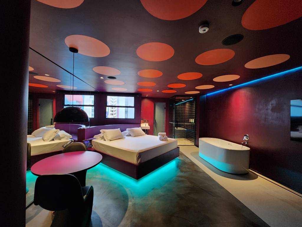 Suítes Andar de Cima-Crédito-Divulgação (13) Imagem de quarto de motel em tom azul e repleto de luzes neon, com cama de casal e chuveiro com janela ao fundo