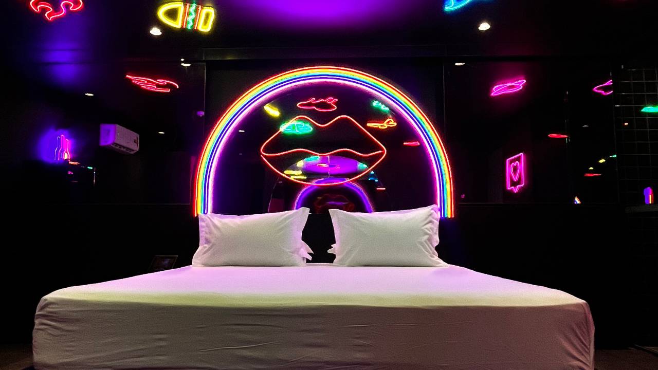 Imagem 3D de quarto de motel repleto de luzes neon e parede preta, com cama de casal no centro