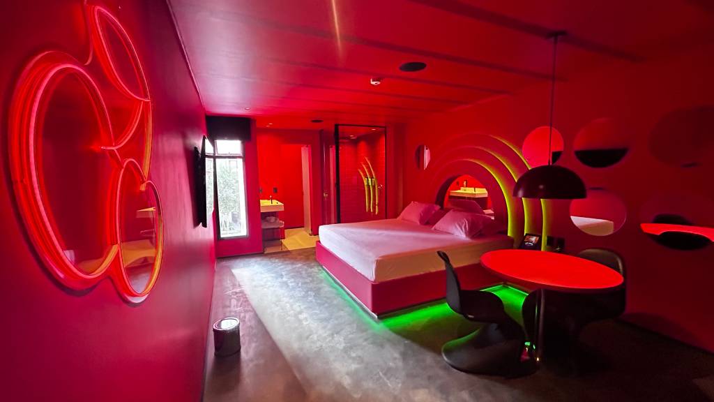Imagem de quarto de motel em tom vermelho e repleto de luzes neon, com cama de casal e chuveiro com janela ao fundo