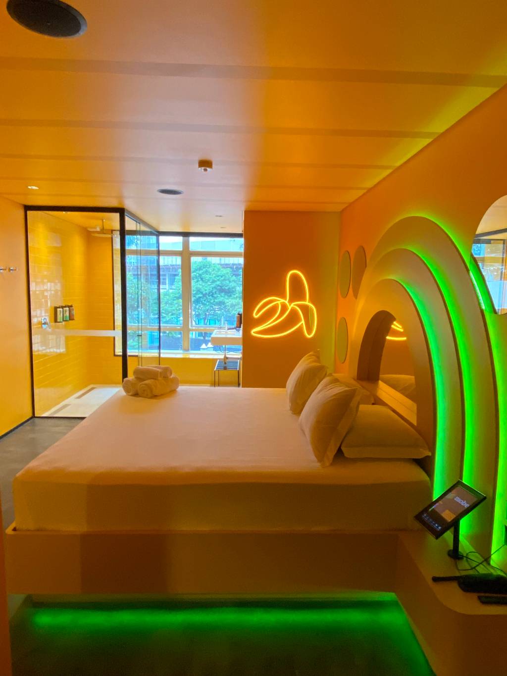 Imagem de quarto de motel em tom amarelo e repleto de luzes neon, com cama de casal e chuveiro com janela ao fundo. Uma das paredes tem um neon em formato de banana.