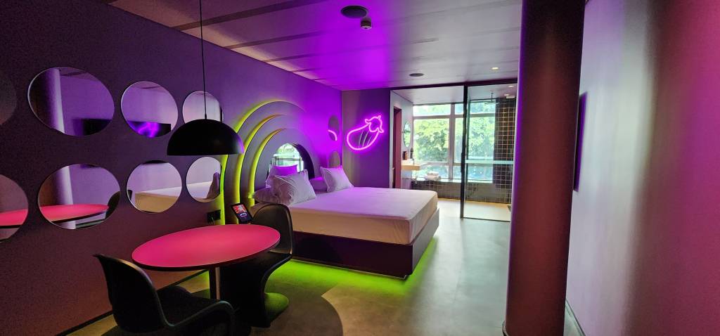 Imagem de quarto de motel em tom roxo e repleto de luzes neon, com cama de casal e chuveiro com janela ao fundo
