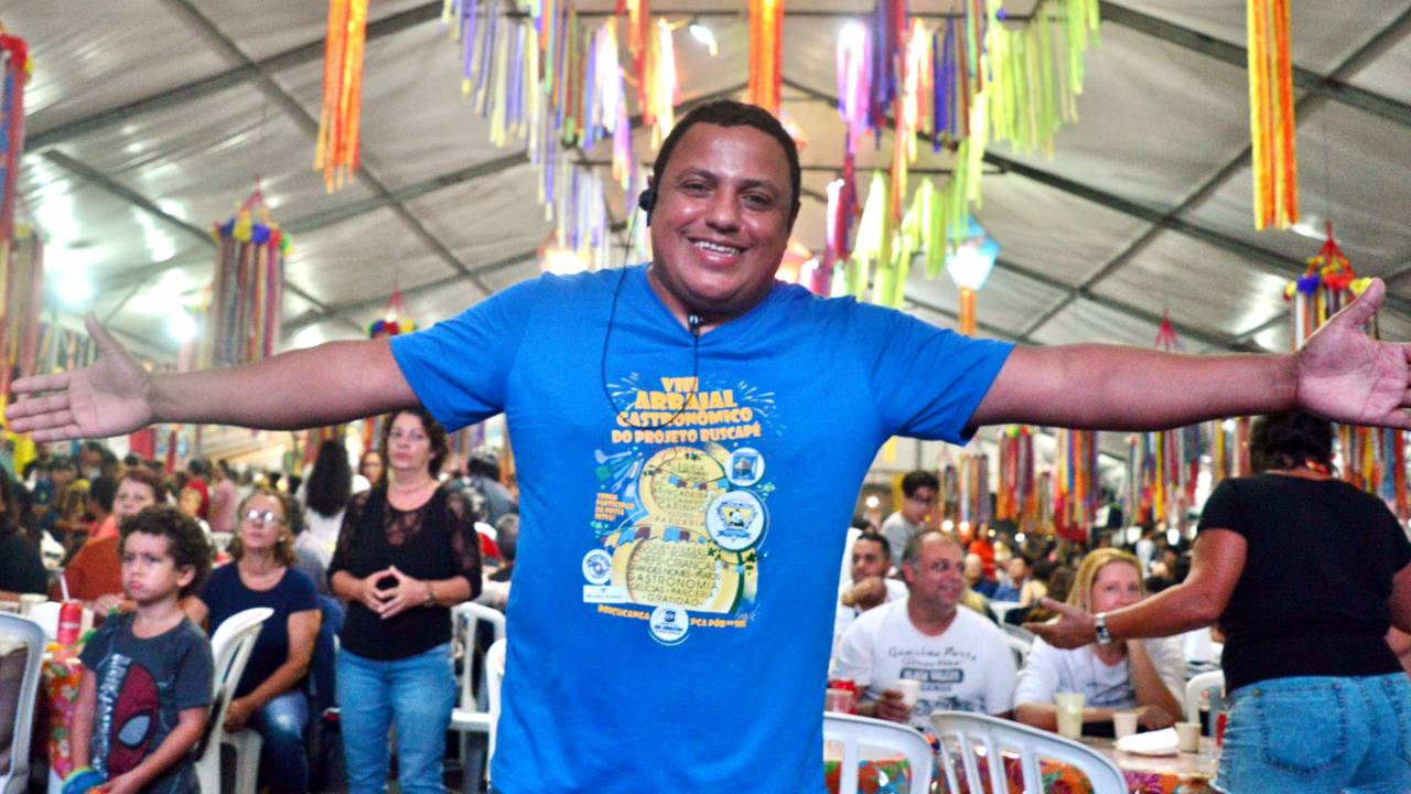 Homem de braços abertos em frente a salão de festa junina.