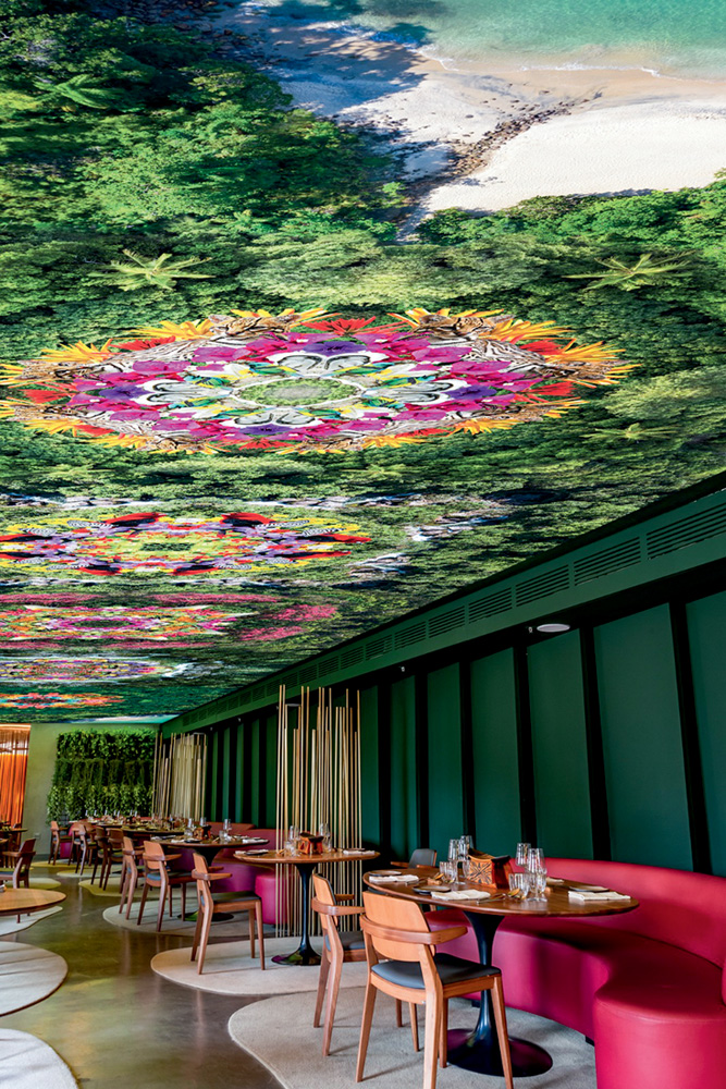 Projeto do salão do restaurante Notiê. Com teto pintado colorido, à direita com mesa de madeira e sofá de couro vermelho.