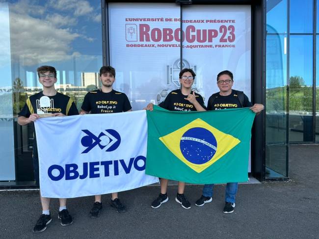 Imagem mostra quatro pessoas, três adolescentes e um adulto, segurando bandeira do Brasil e bandeira do Colégio Objetivo em frente a uma parede de vidro, com cartaz escrito: 