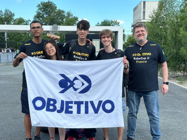 Imagem mostra quatro jovens e um adulto segurando bandeira do colégio Objetivo, sorrindo para foto