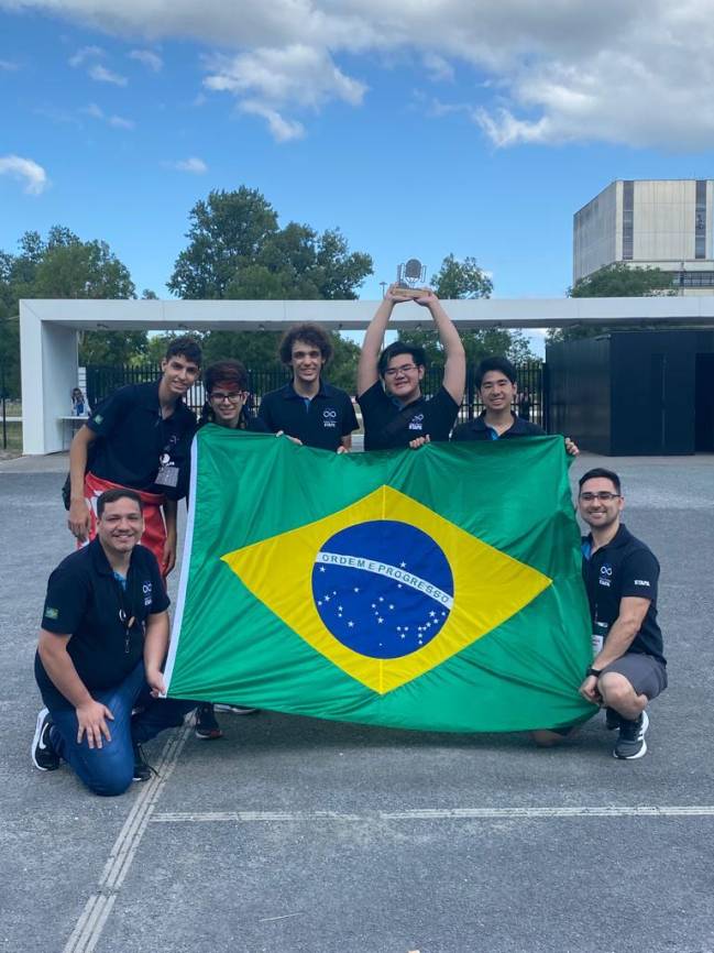 Imagem mostra 7 homens segurando a bandeira do Brasil em ambiente aberto, sorrindo para foto
