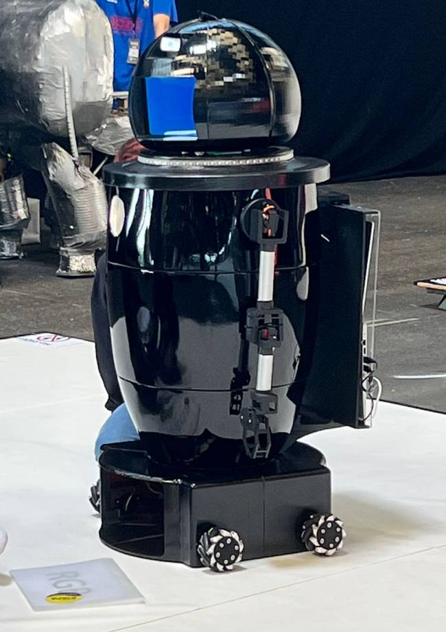 robocup frança Imagem mostra robô composto por três partes. A base é quadrada com rodinhas, o meio se parece com um barril e a cabeça é redonda. O robô está em um palco branco