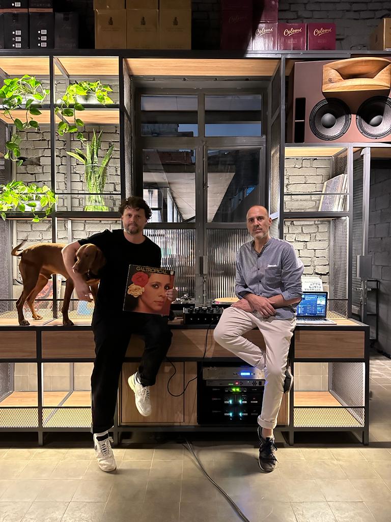 A imagem mostra os dois sócios do bar sentados em uma mesa com equipamentos de som. Leandro Mattiuz à esquerda segura um cachorro com um braço e com o outro um disco de Gal Costa.