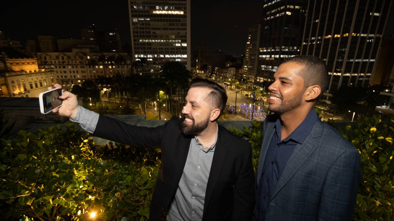 Imagem mostra dois homens tirando selfie em cenário urbano