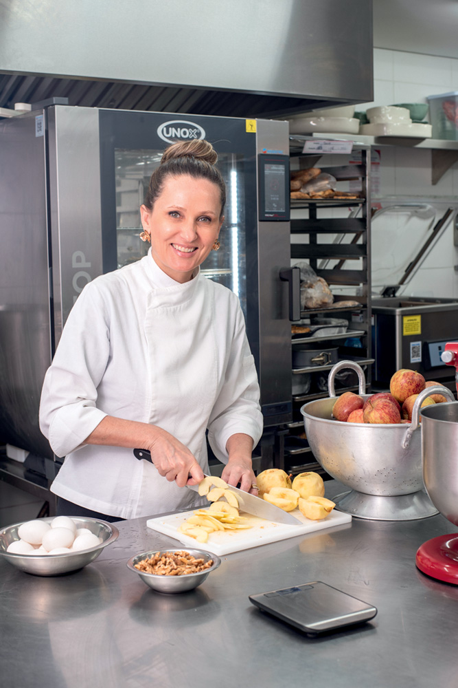 Chef de cozinha loira com cabelo preso em coque e vestida de dólmã cortando pêssegos em cozinha industrial de restaurante ao sorrir para a foto.