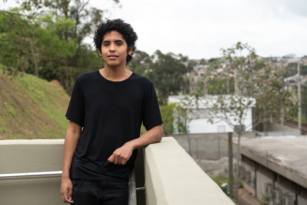 Foto mostra jovem em cima de passarela, apoiado com o cotovelo em sacada, olhando para foto. Paisagem atrás tem árvores e prédios, ao redor de estação de trem