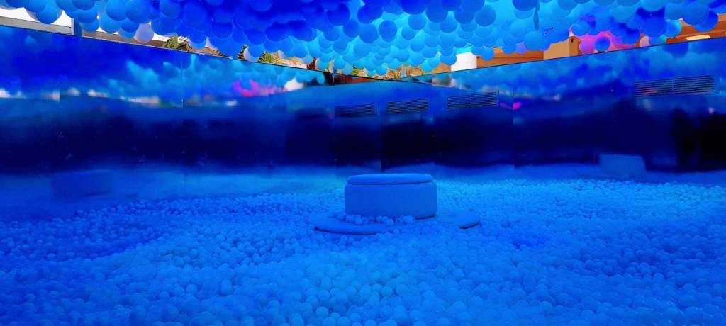 Balloon Experience_1 A imagem mostra uma sala ampla com uma piscina de bolinhas coloridas e balões pendurados no teto.