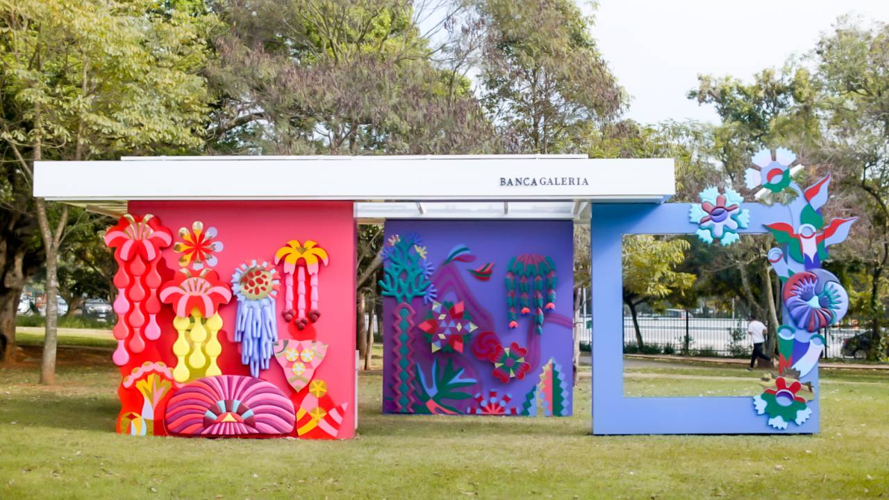 Imagem em gramado exibe banca aberta com duas paredes coloridas e adornadas por figuras abstratas. Uma é vermelha, outra azul.