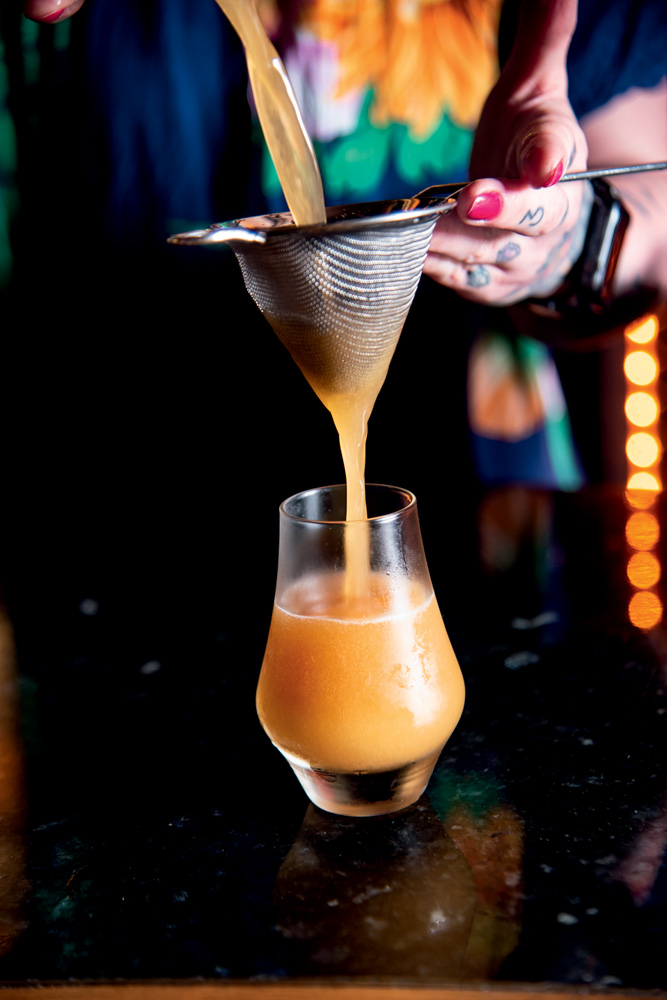 A imagem mostra um copo de vidro com um drinque cor caramelo dentro. Uma pessoa despeja o drinque no copo com uma peneira.