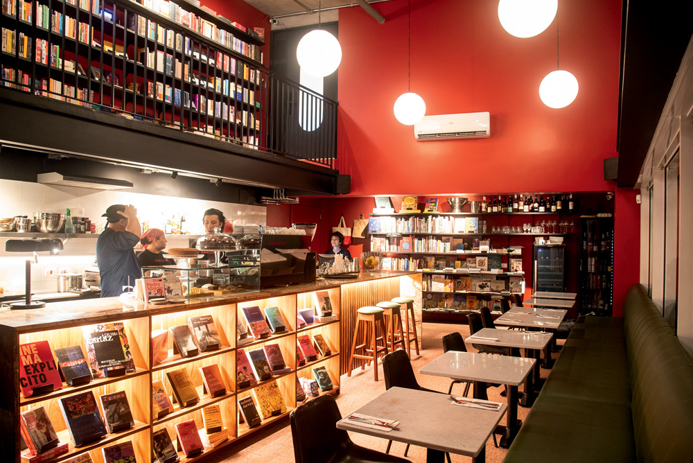 Ambiente interno do bar, com mesas e cadeiras à esquerda, e prateleiras de livros à direita e ao fundo. As paredes são vermelhas.