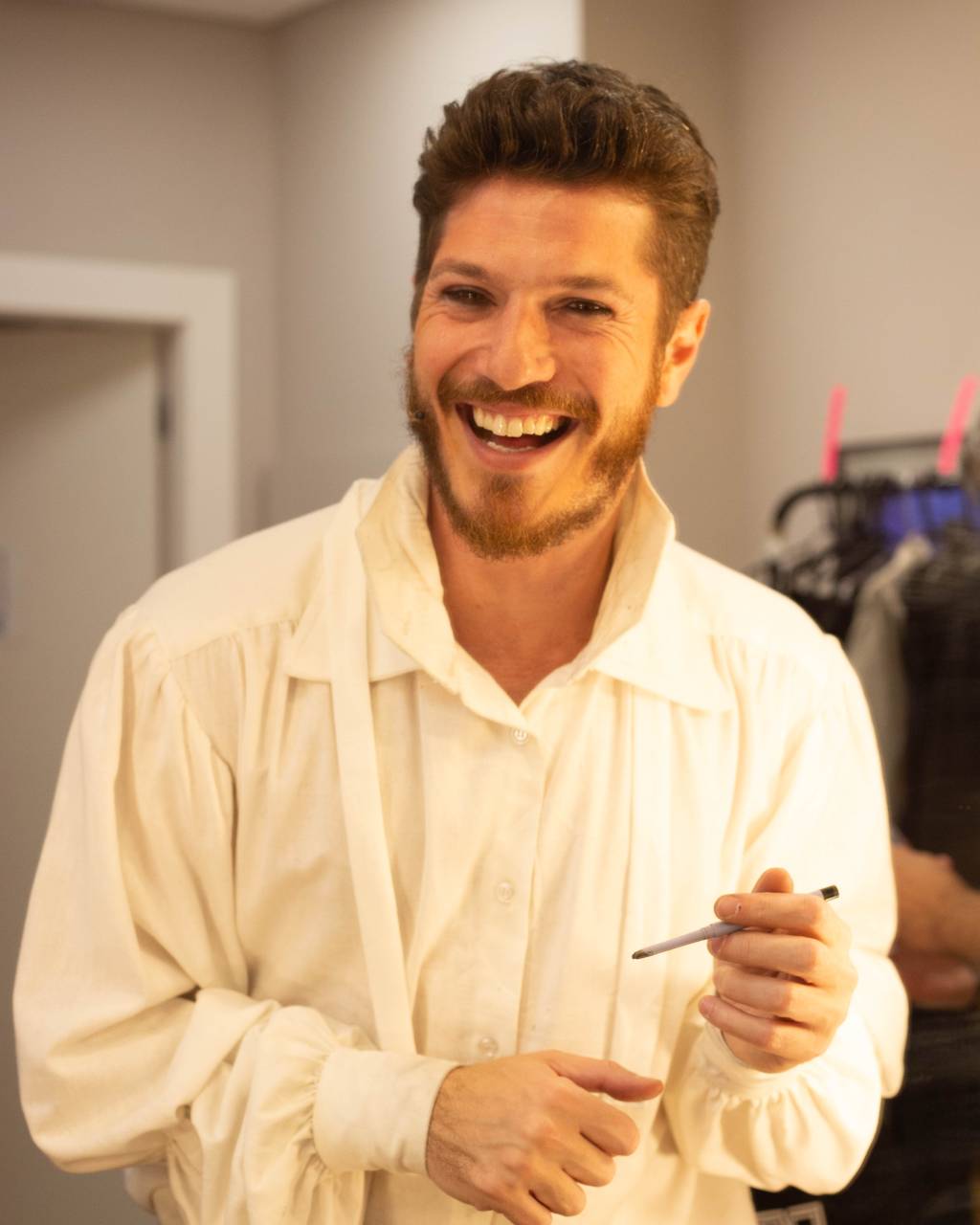 Homem loiro branco com camisa branca sorri em camarim. Segura uma caneta.