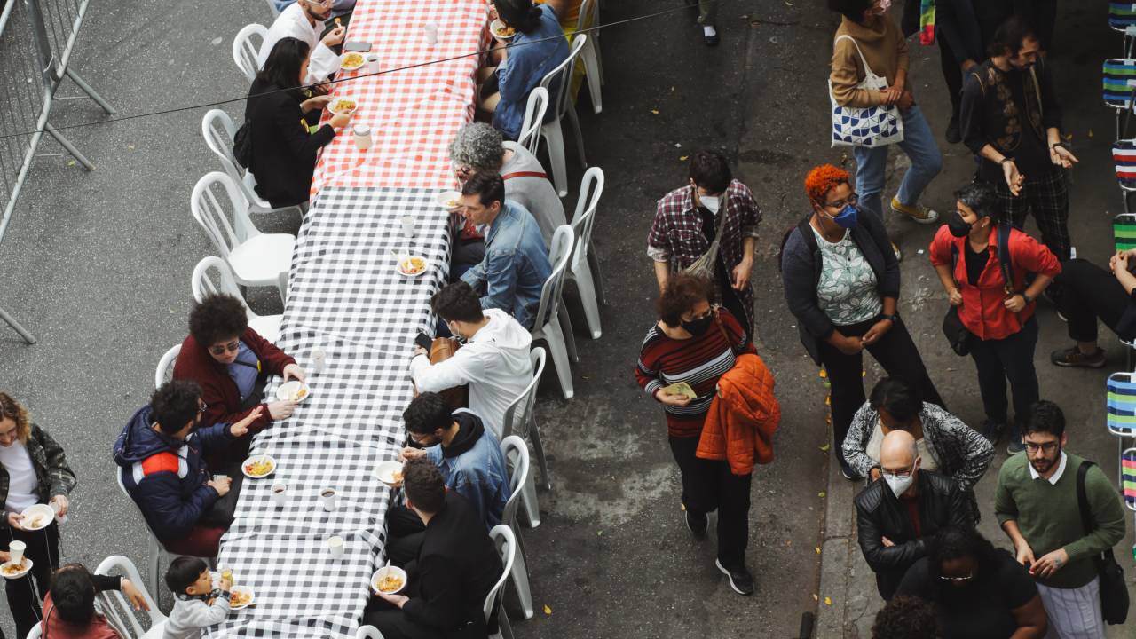 Imagem mostra várias pessoas sentadas em grande mesa no Bom Retiro, almoçando
