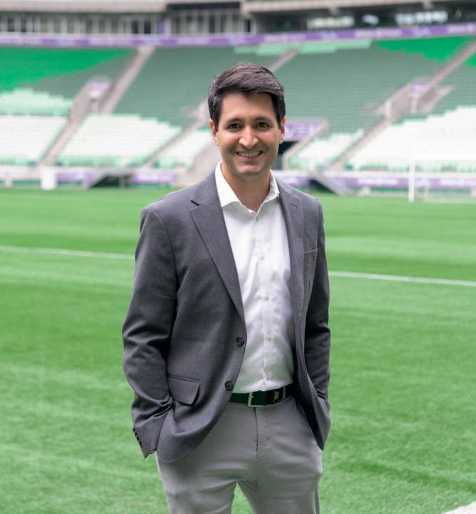 Imagem mostra Cláudio Macedo em campo de futebol, usando blazer, camisa e calça social. Ele sorri para a foto em dia ensolarado