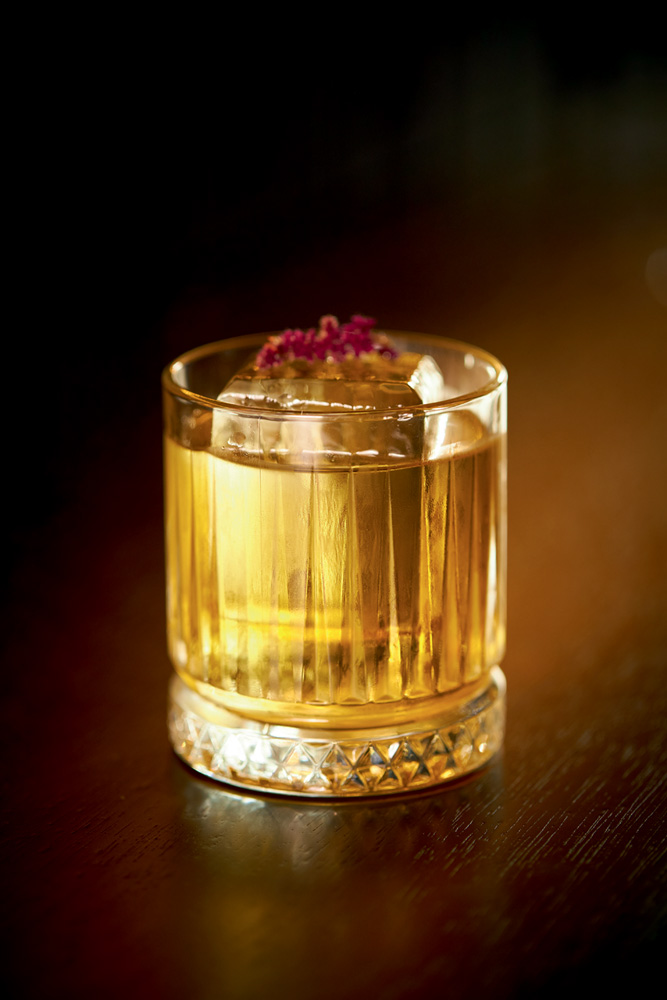 Drinque amarelado em copo baixo com gelão coberto por flor roxa sobre mesa de madeira.