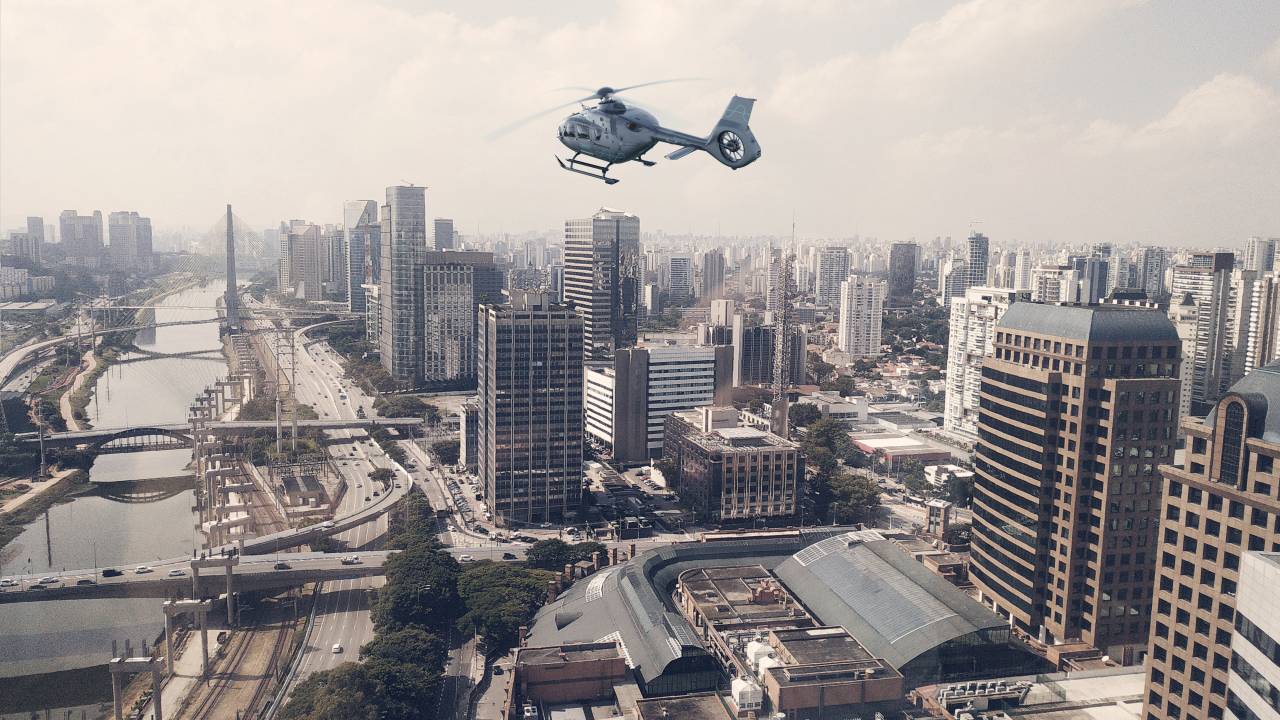 Foto exibe helicóptero sobrevoando área repleta de prédios e avenidas.