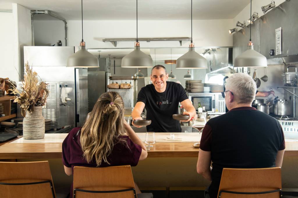 Fame Chef de cozinha atrás de balcão entregando prato para dois clientes.