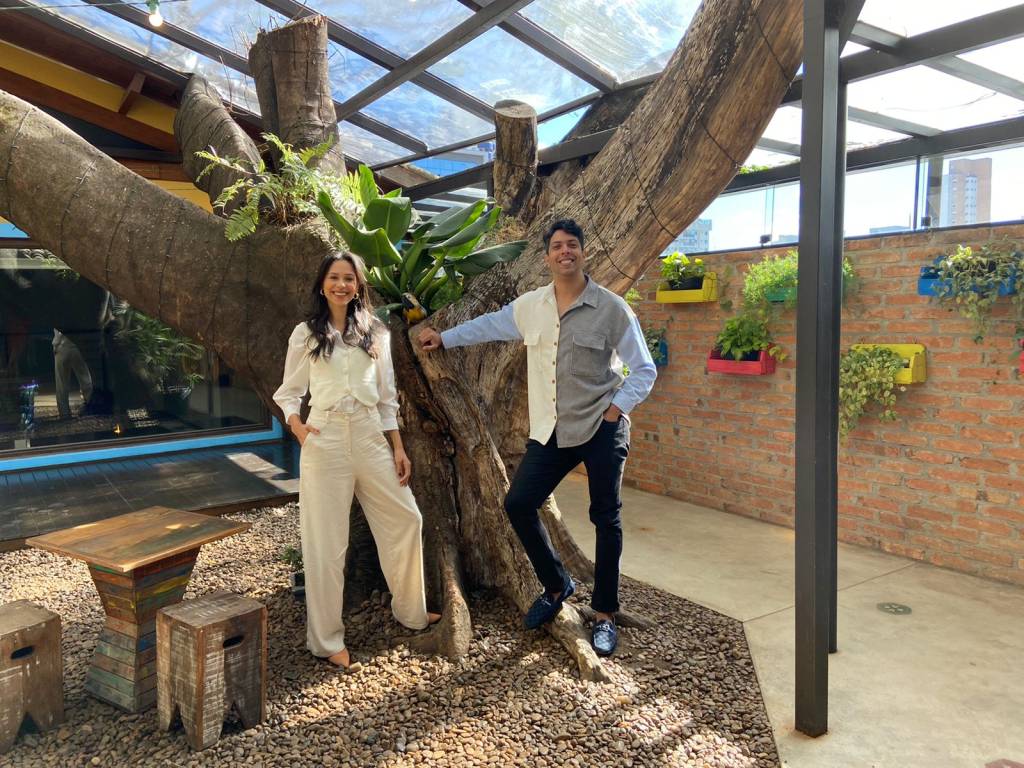 Moça ruiva sorri em cenário com plantas penduradas em fundo branco. Veste vestido preto