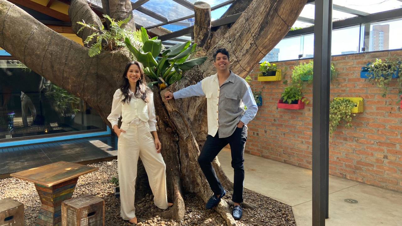 Casal posa em frente a grande árvore em quintal. Ambos têm a pele parda e aparecem de pé. Ela veste roupa branca e ele uma calça preta com camisa metade branca, metade azul.