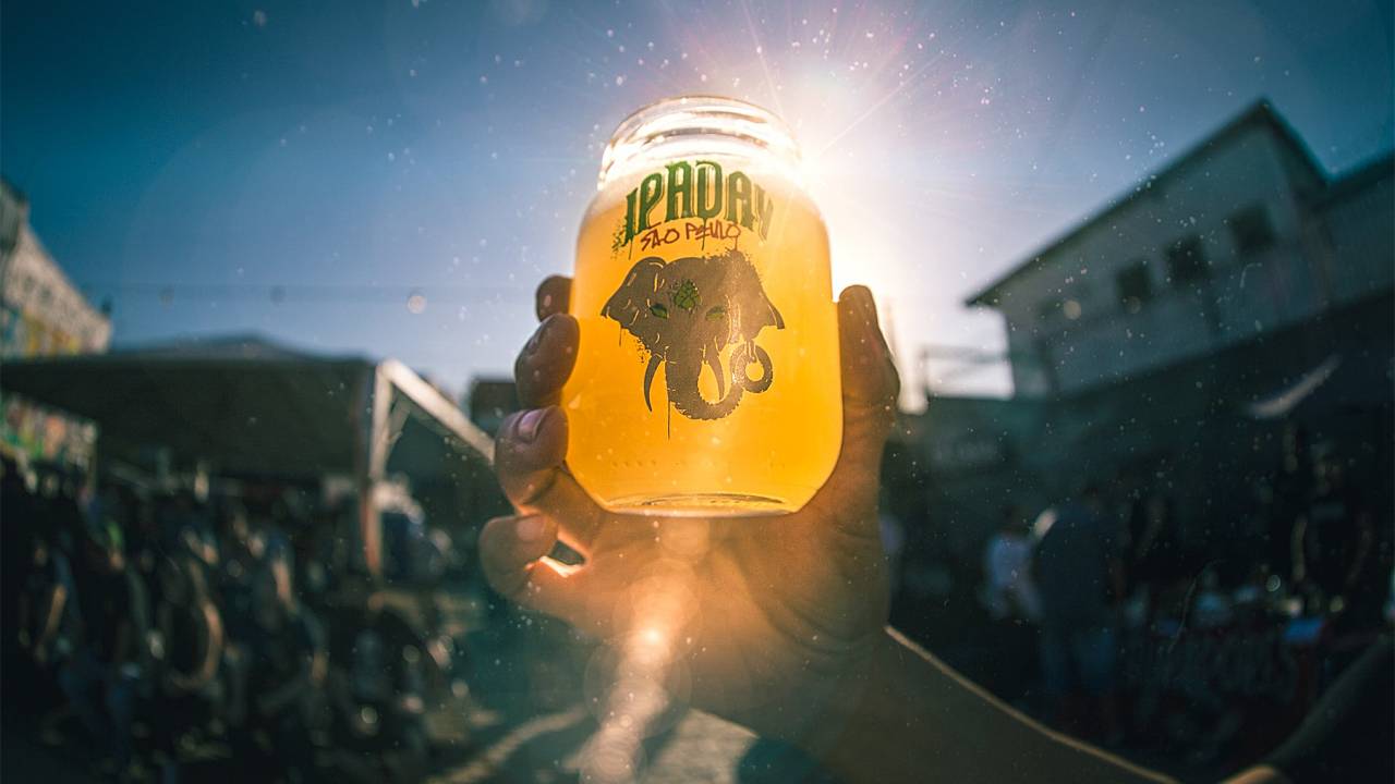 A imagem mostra uma mão segurando um copo de vidro com cerveja contra a luz do sol.