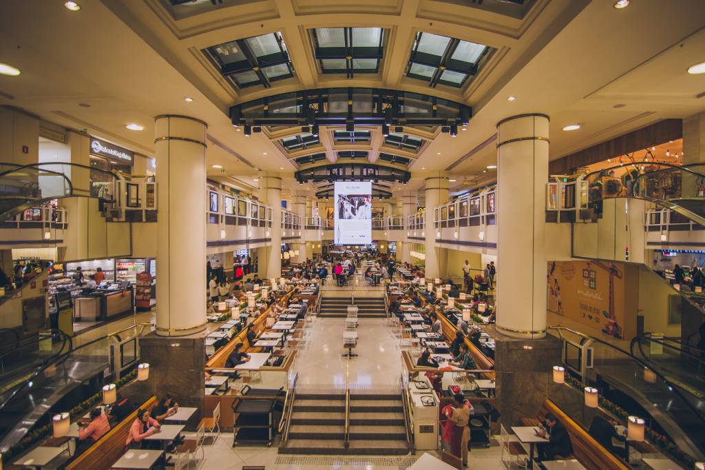 Foto exibe área central de shopping com teto envidraçado e colunas brancas.