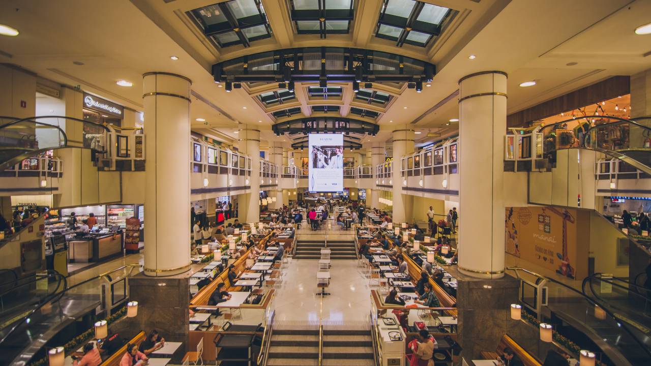 Foto exibe área central de shopping com teto envidraçado e colunas brancas.