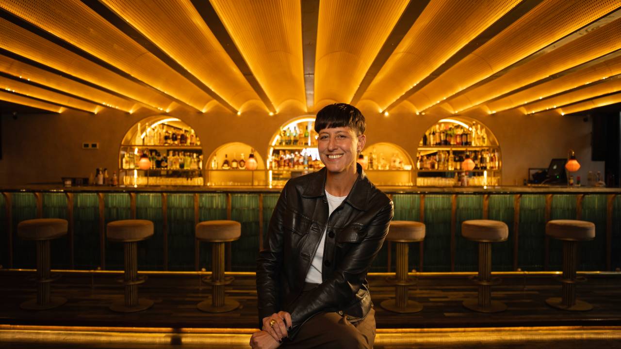 A imagem mostra a bartender sentada em um banco e sorrindo para a foto. Ela usa uma jaqueta preta e calça marrom. Ao fundo aparece a mesa do bar com banquinhos na frente e bebidas atrás.
